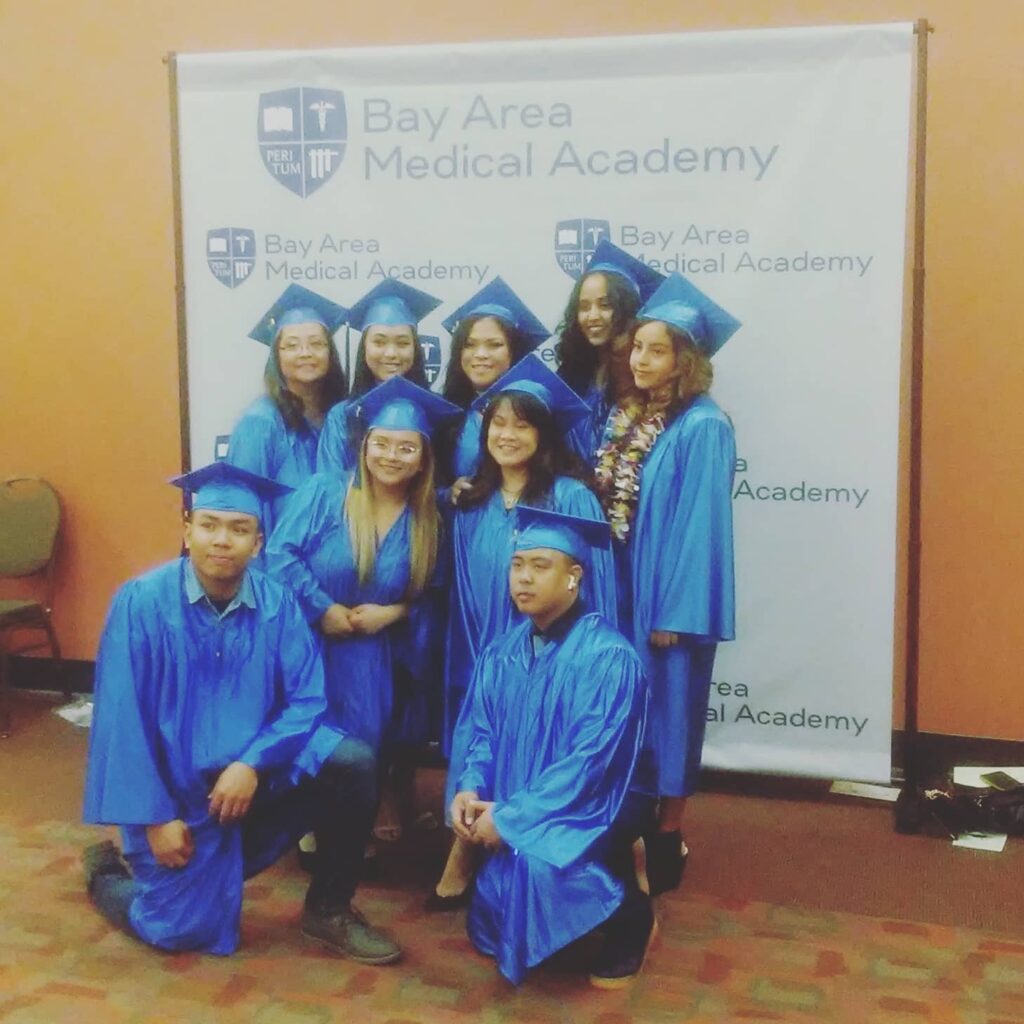 A Group of Graduates pose cheerfully in front of the Bay Area Medical Academy Banner.